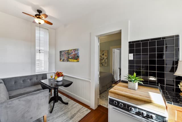 A dining area featuring a grey tufted settee and a dark wood pedestal table, situated next to a kitchen with black subway tile and a white stove. An open doorway reveals a glimpse of a bedroom beyond.