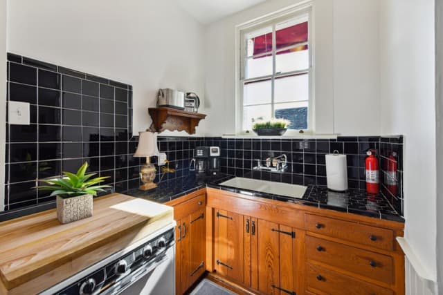 A kitchen featuring warm wood cabinets and a black tile countertop and backsplash. A window above the sink lets in natural light, and a small wooden cutting board sits atop a white stove in the foreground.