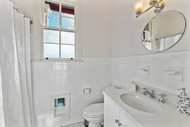 A clean, white bathroom featuring a white vanity with an oval sink, a toilet, and a window with a white frame. The lower half of the walls are finished with white vertical paneling, and a small round mirror is mounted above the sink.