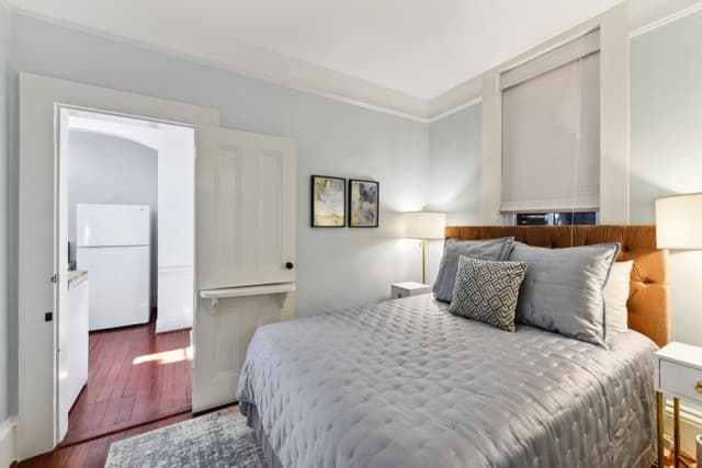 A bedroom with a light grey quilted bedspread and a brown tufted headboard. An open white door leads to a kitchen area where a white refrigerator is visible on dark wood floors. Two small framed pictures hang on the light blue wall above the bed.