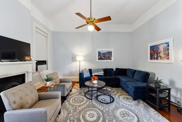 A spacious living room featuring a navy blue sectional sofa and two grey armchairs arranged around a circular coffee table on a patterned rug. The room has light blue walls, a white fireplace mantle with a television above it, and a wooden ceiling fan.