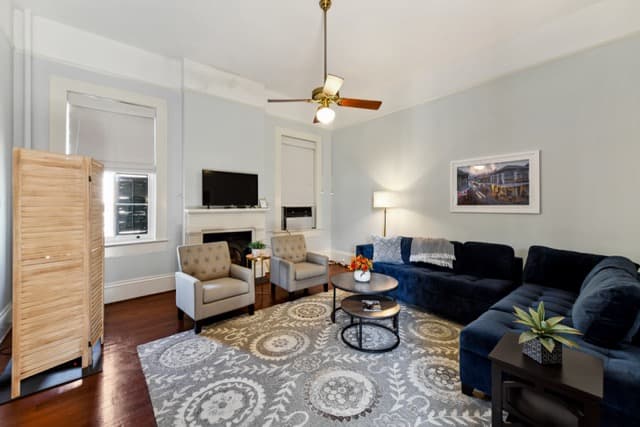 A wide-angle view of a living room featuring a navy blue sectional sofa, two grey tufted armchairs, and a circular nested coffee table on a grey patterned rug. The room includes a fireplace with a TV mounted above it, a brass ceiling fan, and a light wood folding screen to the left.