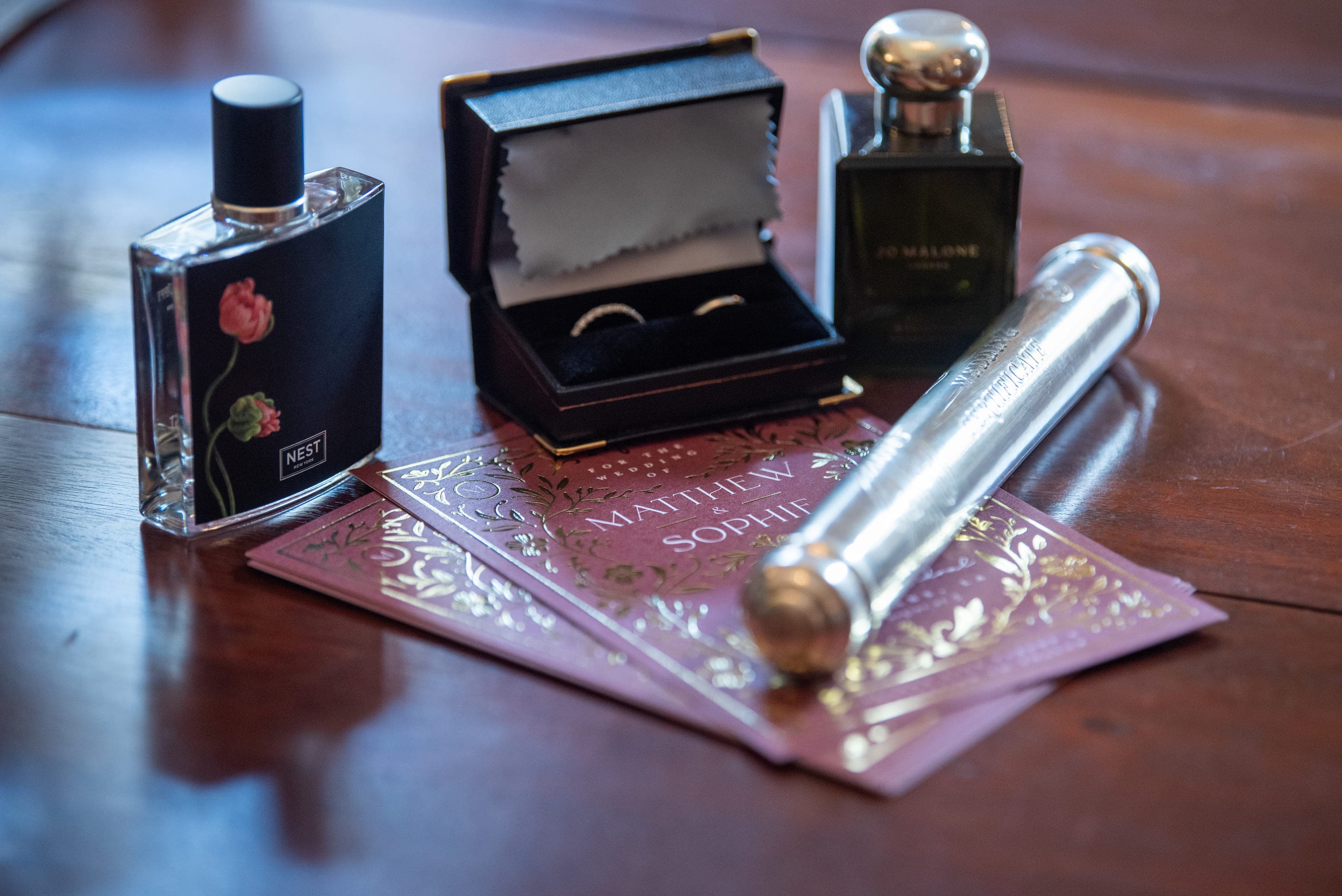 A close-up of a perfume bottle, a ring box containing rings, a second perfume bottle, and decorative invitation cards arranged on a wooden surface.