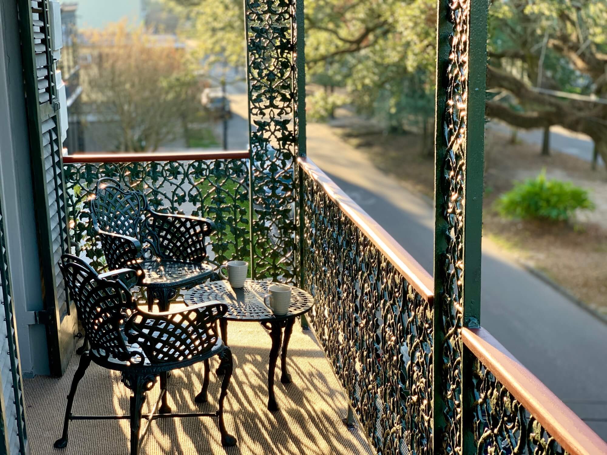 A sunlit balcony with wrought-iron chairs and tables, overlooking a tree-lined street.