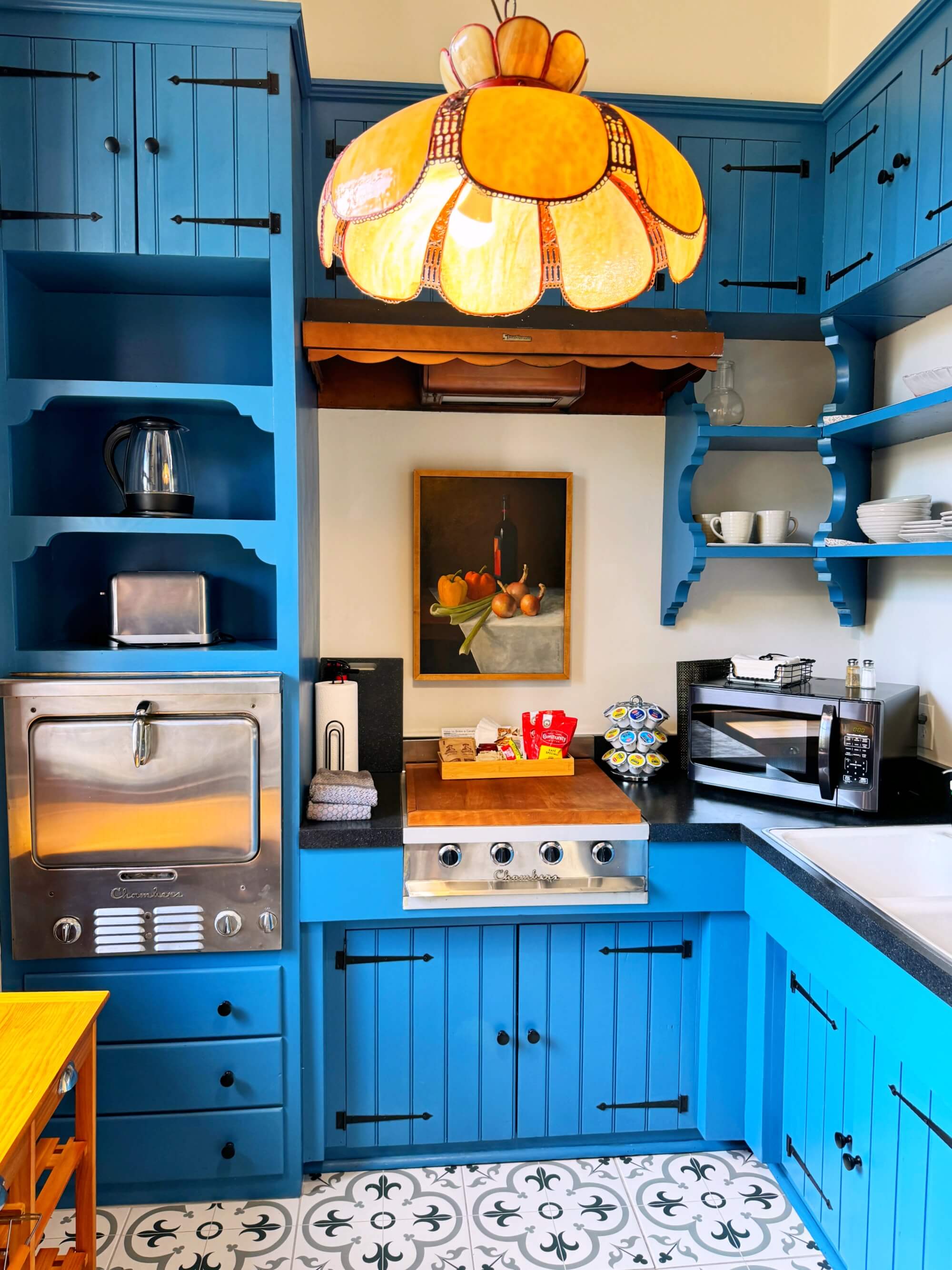 A brightly colored blue kitchen featuring open shelves, a vintage-style lamp, and a painting of fruits.