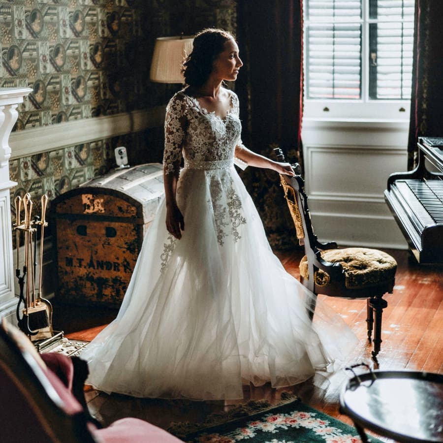 A bride in a lace wedding gown stands in a beautifully decorated room, gazing thoughtfully out a window.