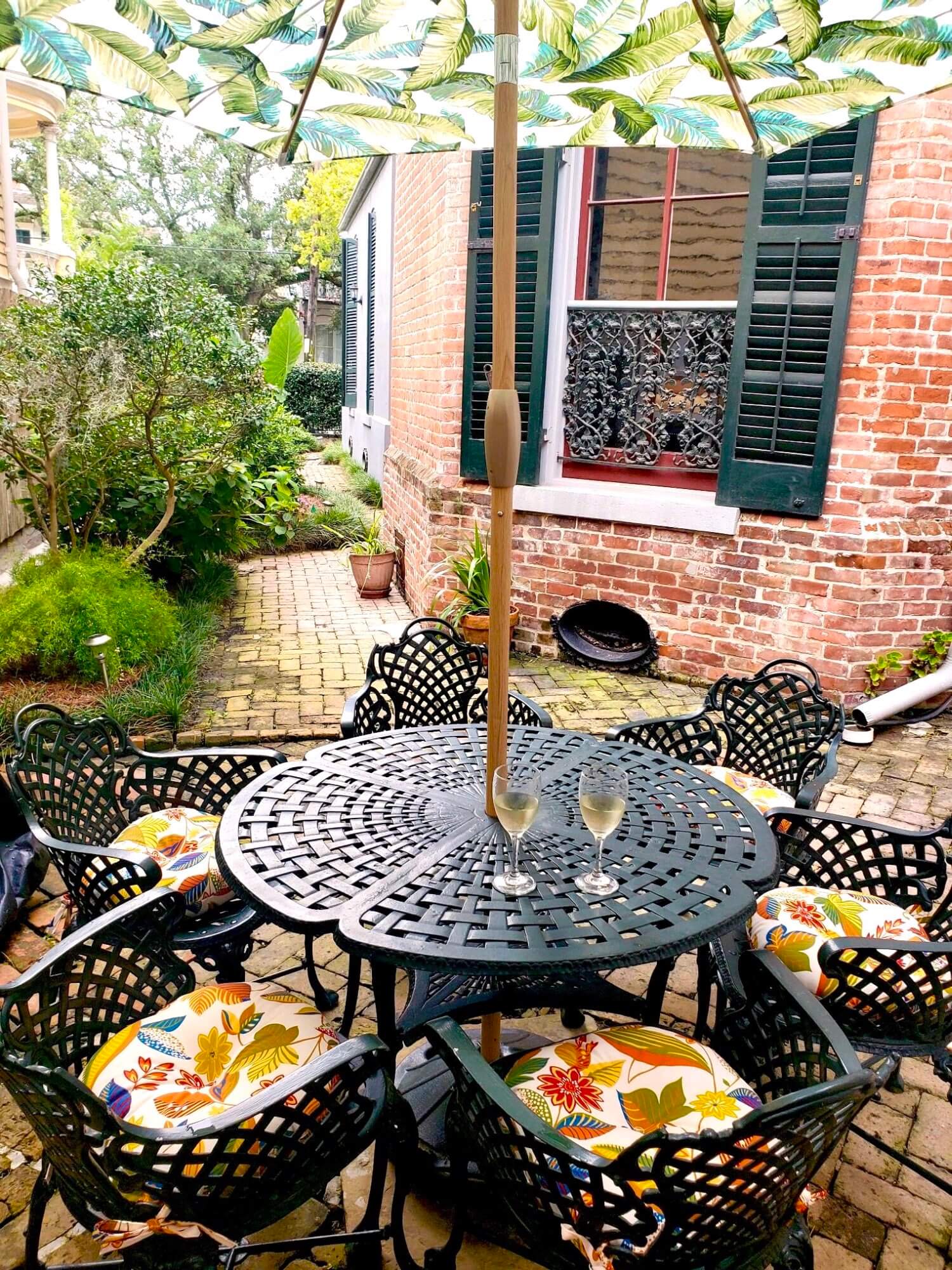 A cozy outdoor dining area with a table, patterned umbrella, and cushioned chairs surrounded by greenery.
