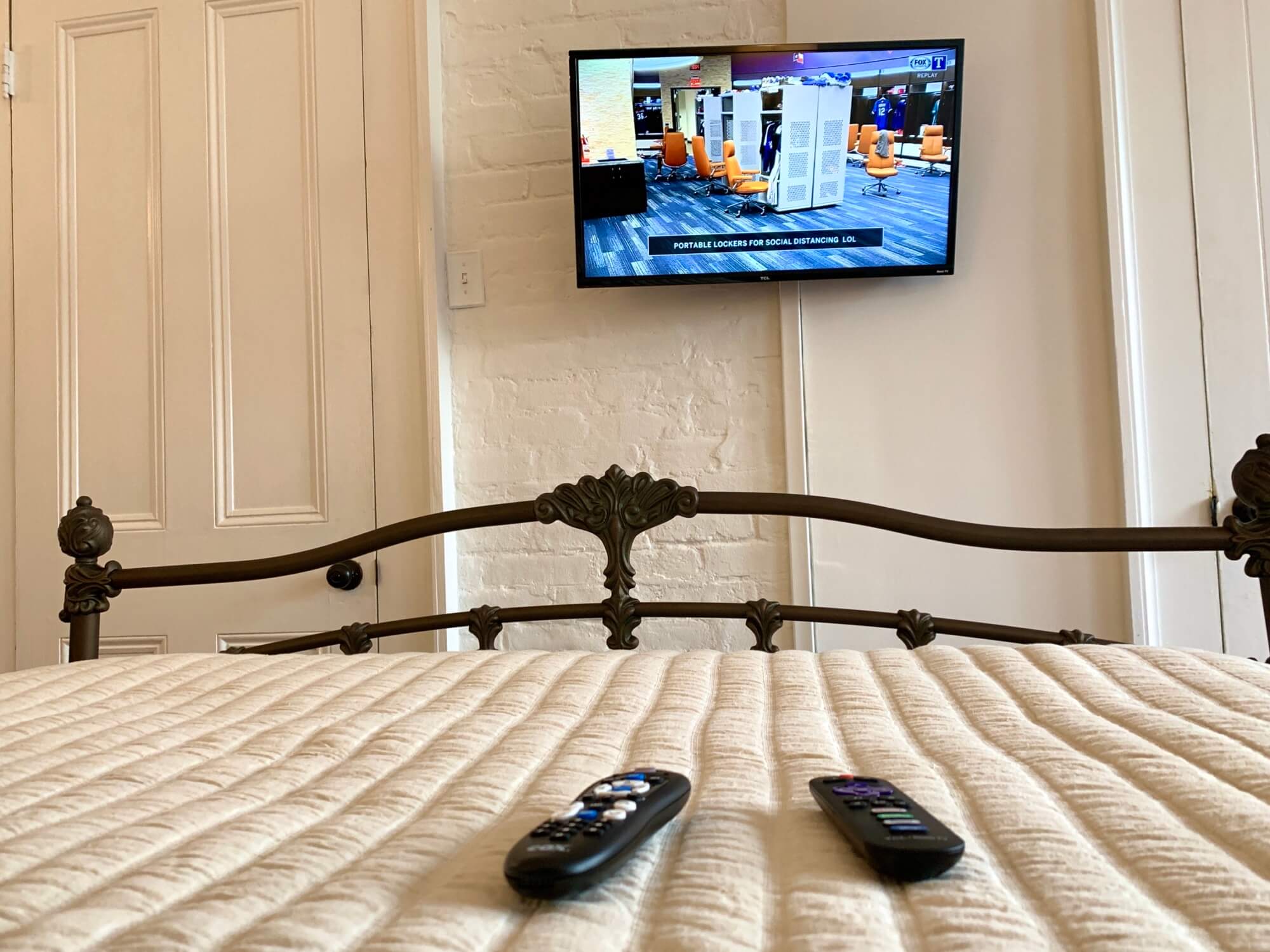 A view from a bed with two remote controls and a television showing a room with orange chairs in the background.