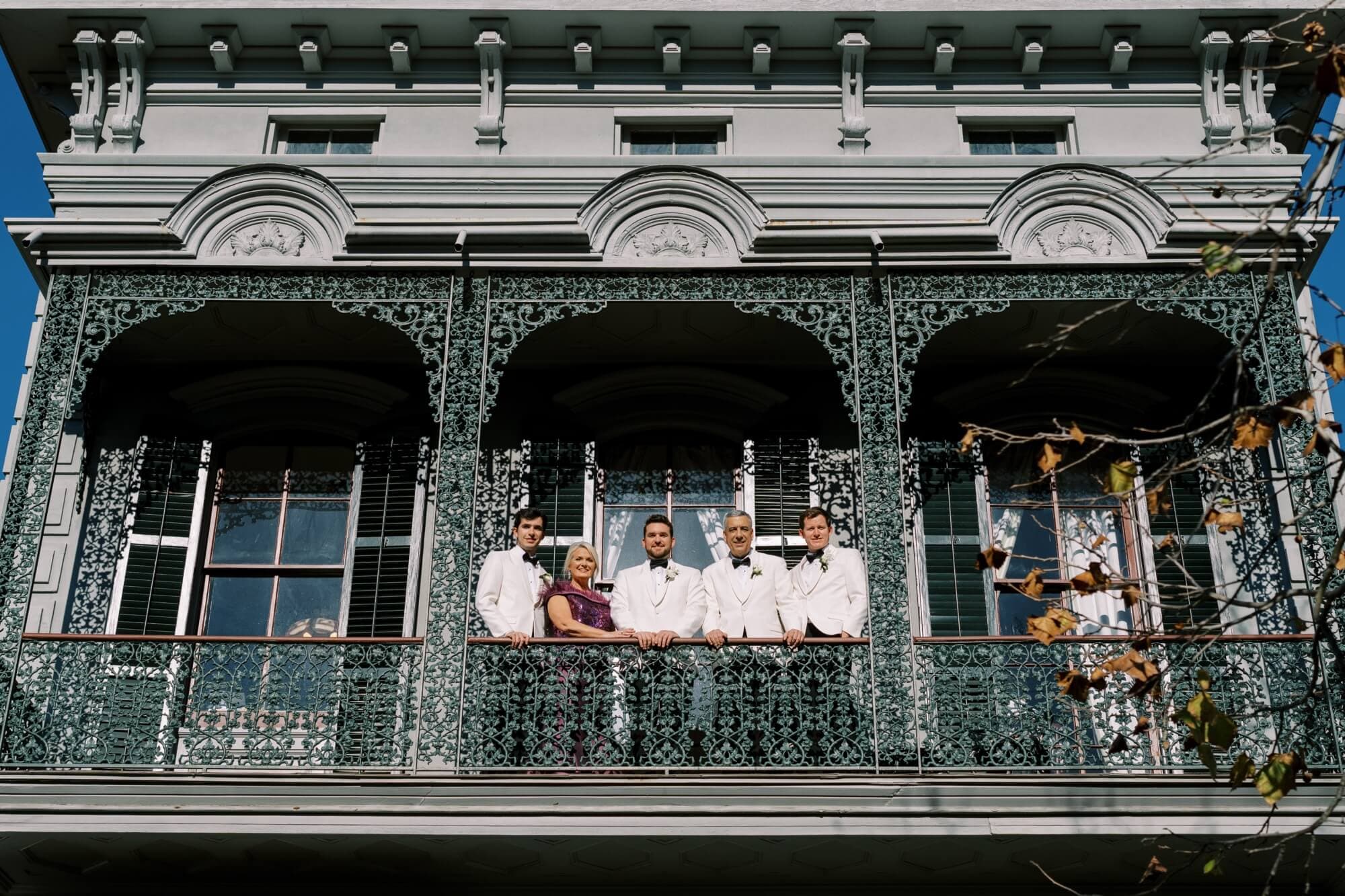 A group of five people in formal attire stands on a decorative balcony with intricate ironwork.