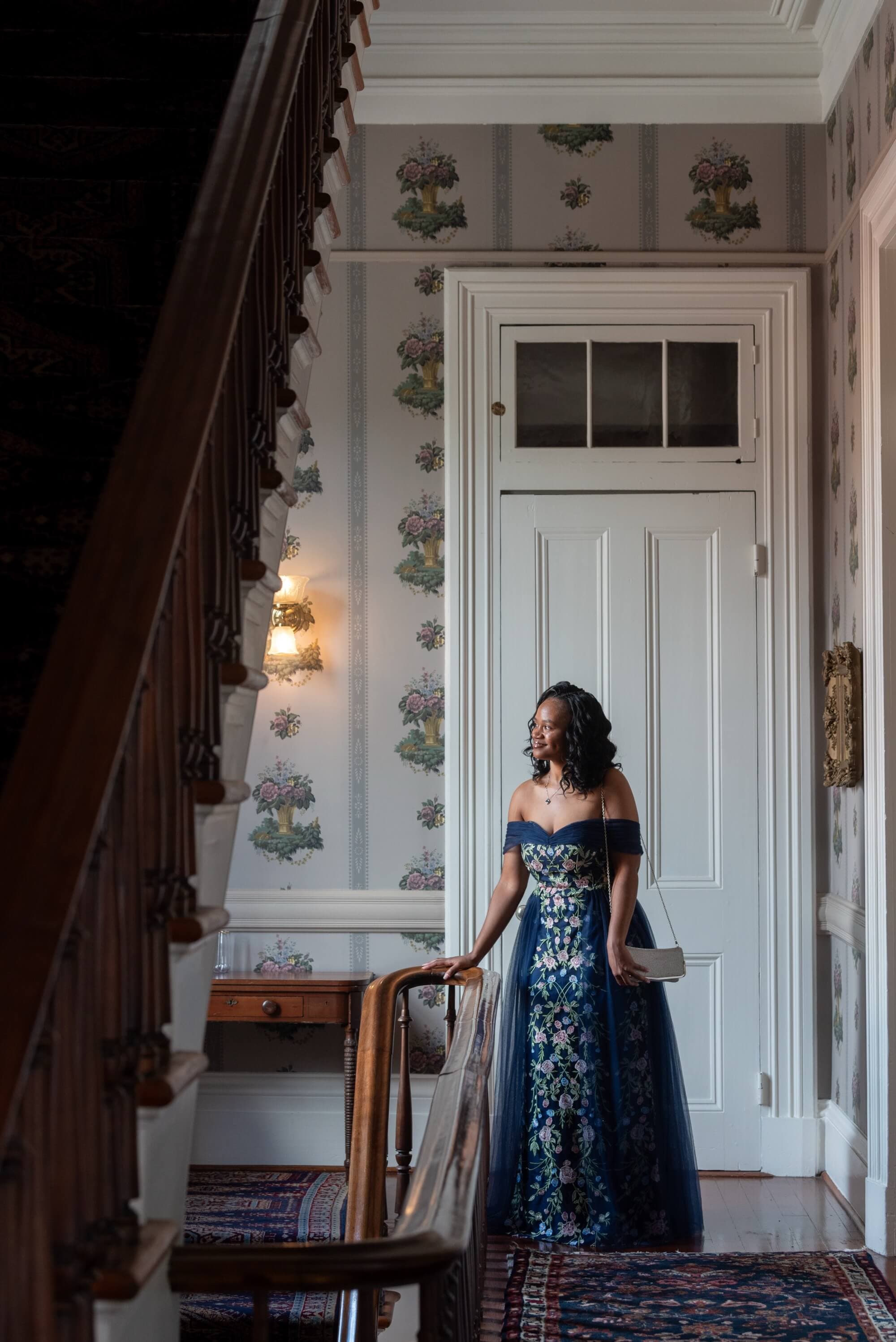 A woman in an elegant blue dress stands by a wooden staircase in a vintage, floral-decorated hallway.