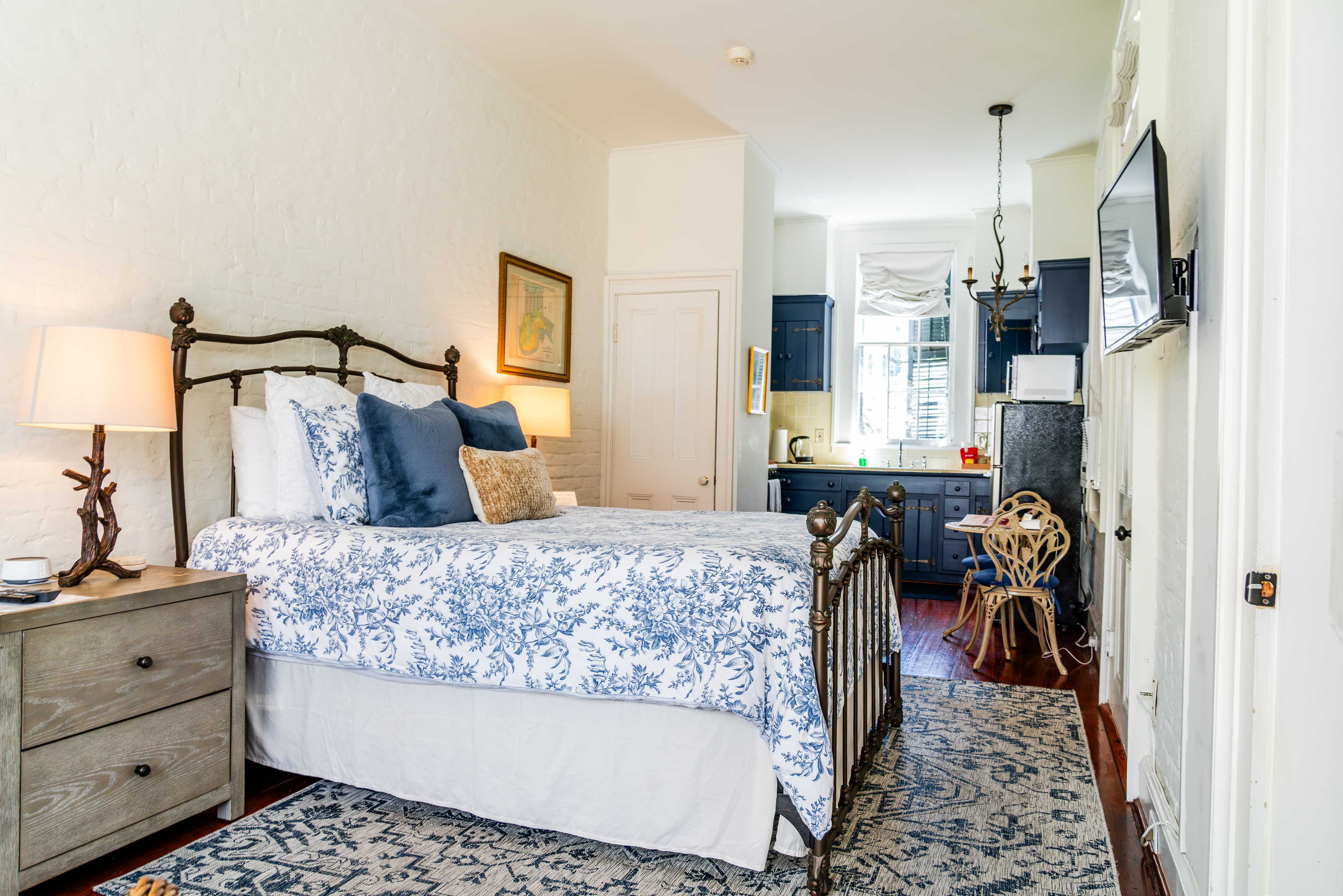 A cozy bedroom featuring a patterned bedspread, bedside table, and kitchenette in the background.