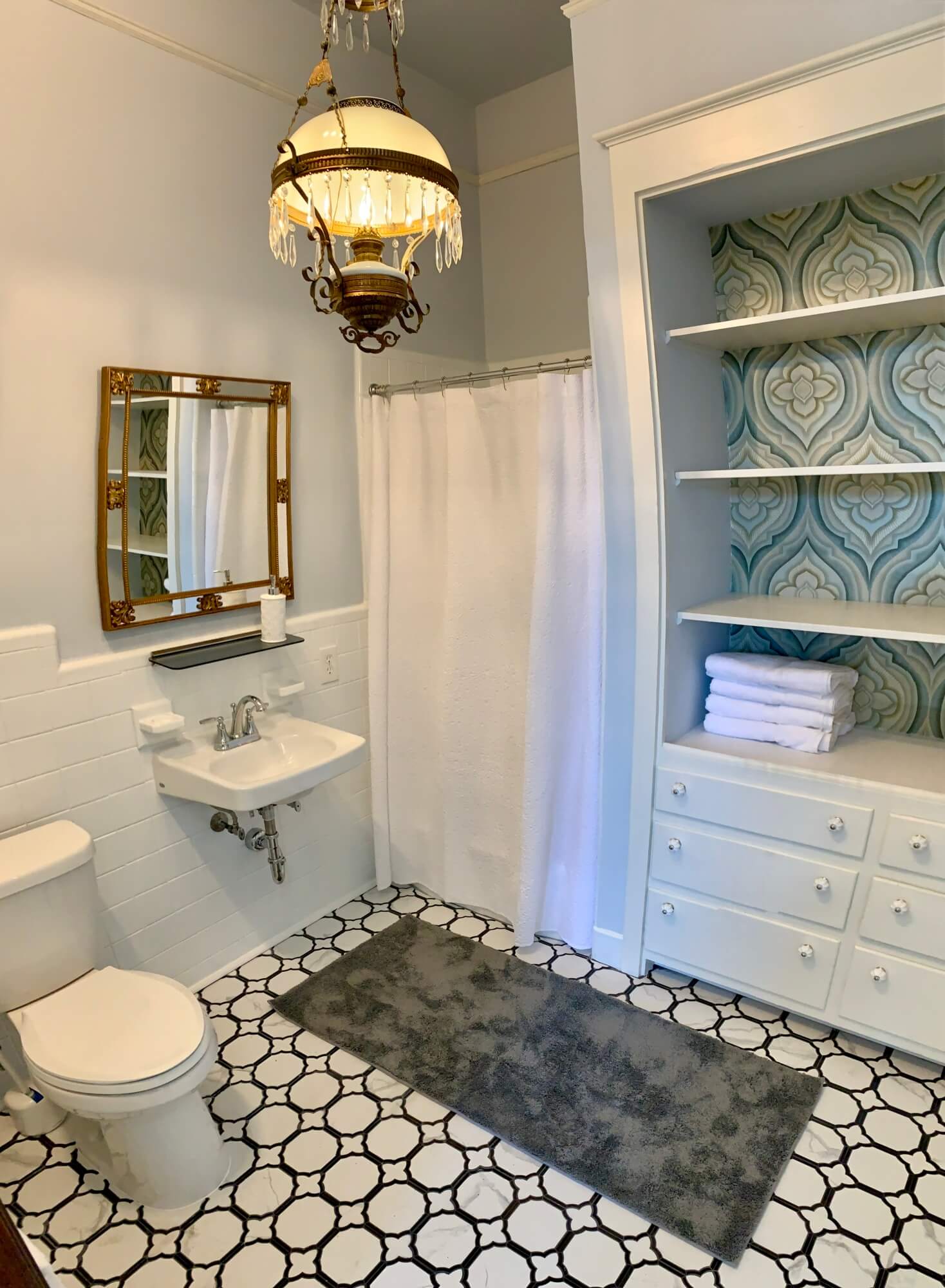 A modern bathroom featuring a decorative chandelier, a shower curtain, a sink, and shelves with neatly stacked towels.