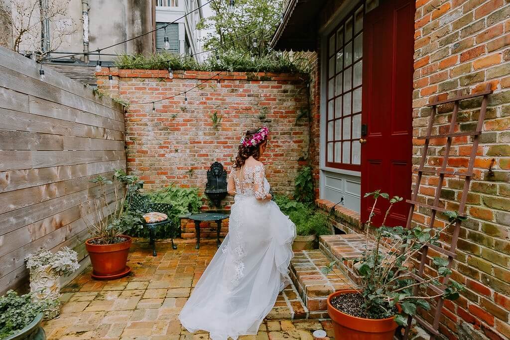 A bride in a flowing white gown and floral crown stands in a charming brick courtyard with potted plants.