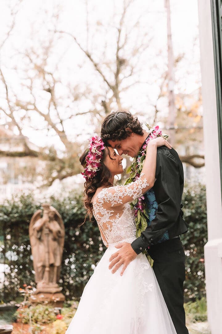 A smiling couple in wedding attire share a romantic embrace outdoors, surrounded by greenery.