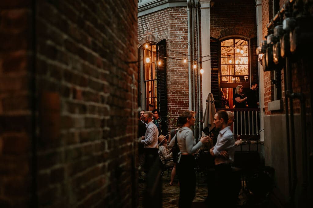 A lively gathering of people in a brick courtyard, illuminated by string lights and glowing windows.