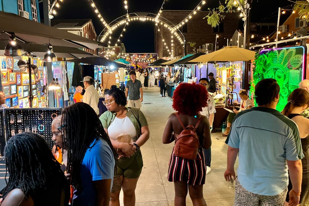 A vibrant night market scene filled with people browsing art stalls under string lights.
