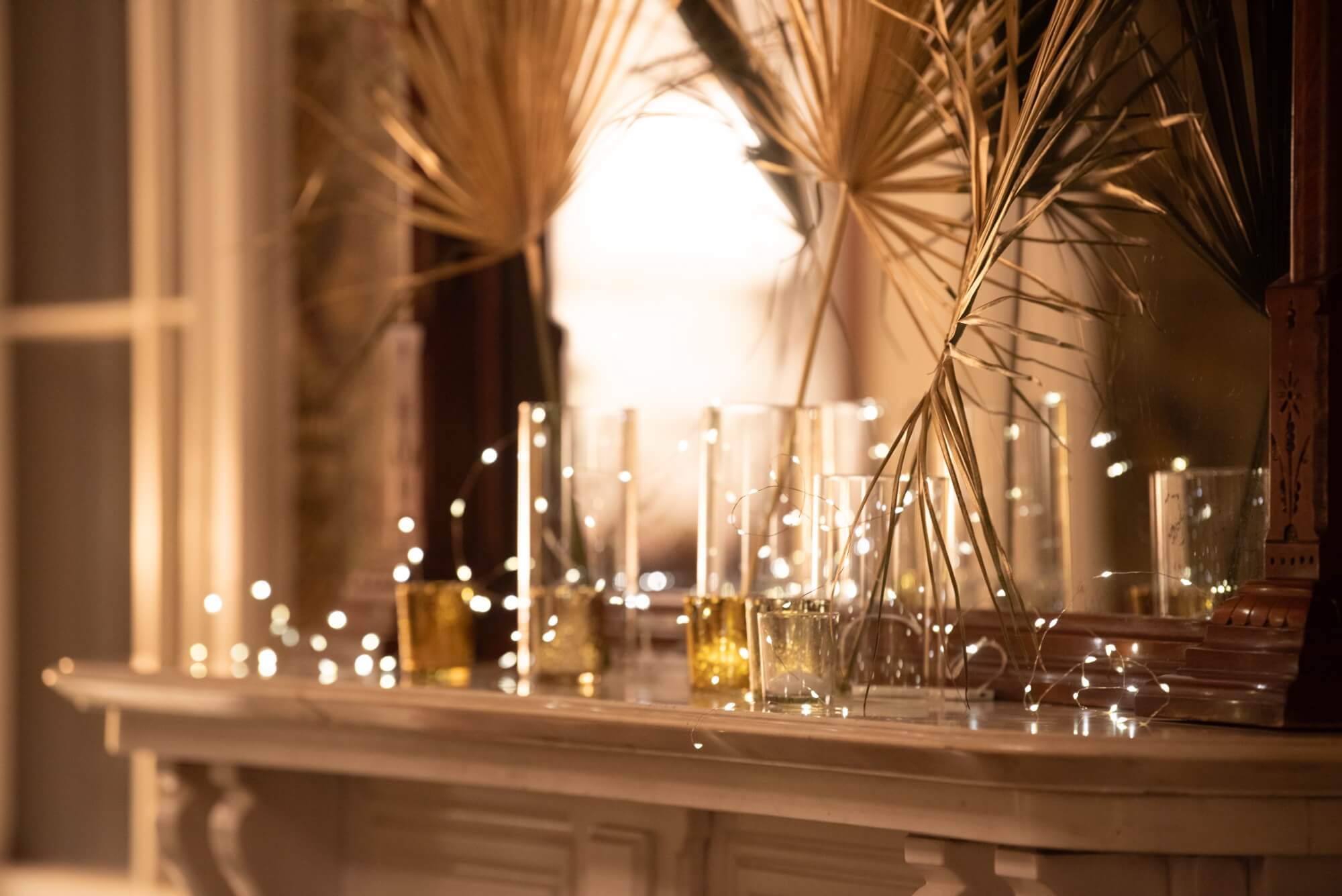 A softly lit mantel displays glass vases and twinkling fairy lights, accented by dried palm fronds.
