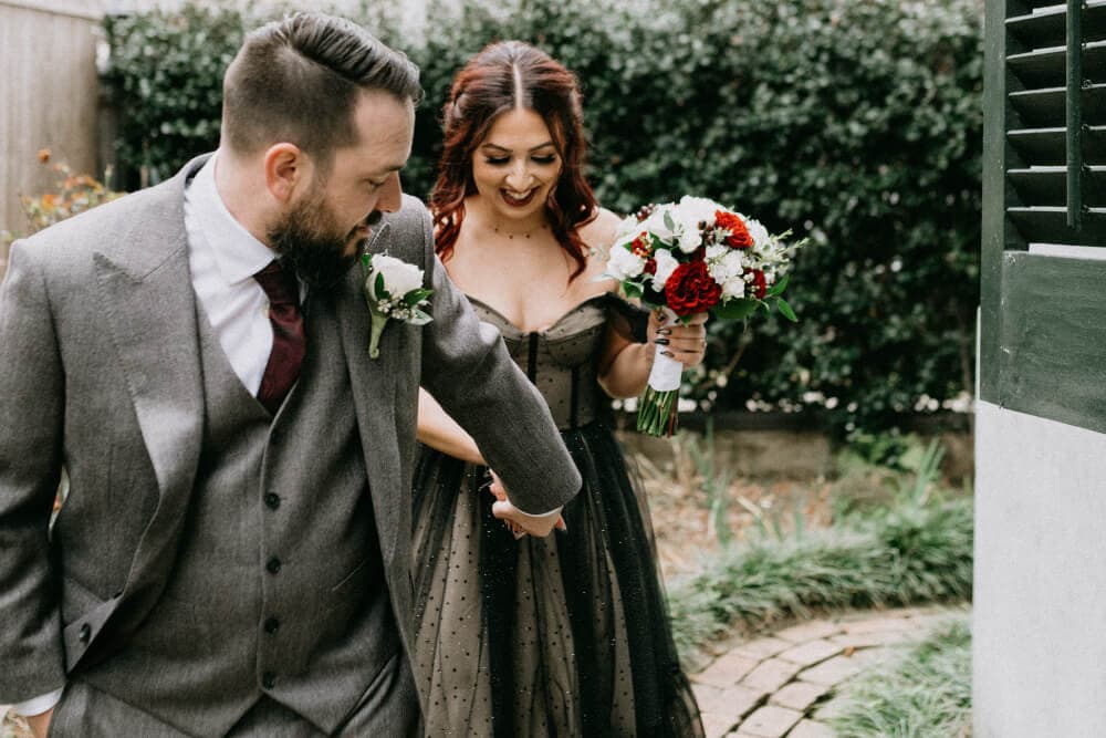 A smiling couple walks hand-in-hand in a garden, with the woman holding a bouquet of flowers.