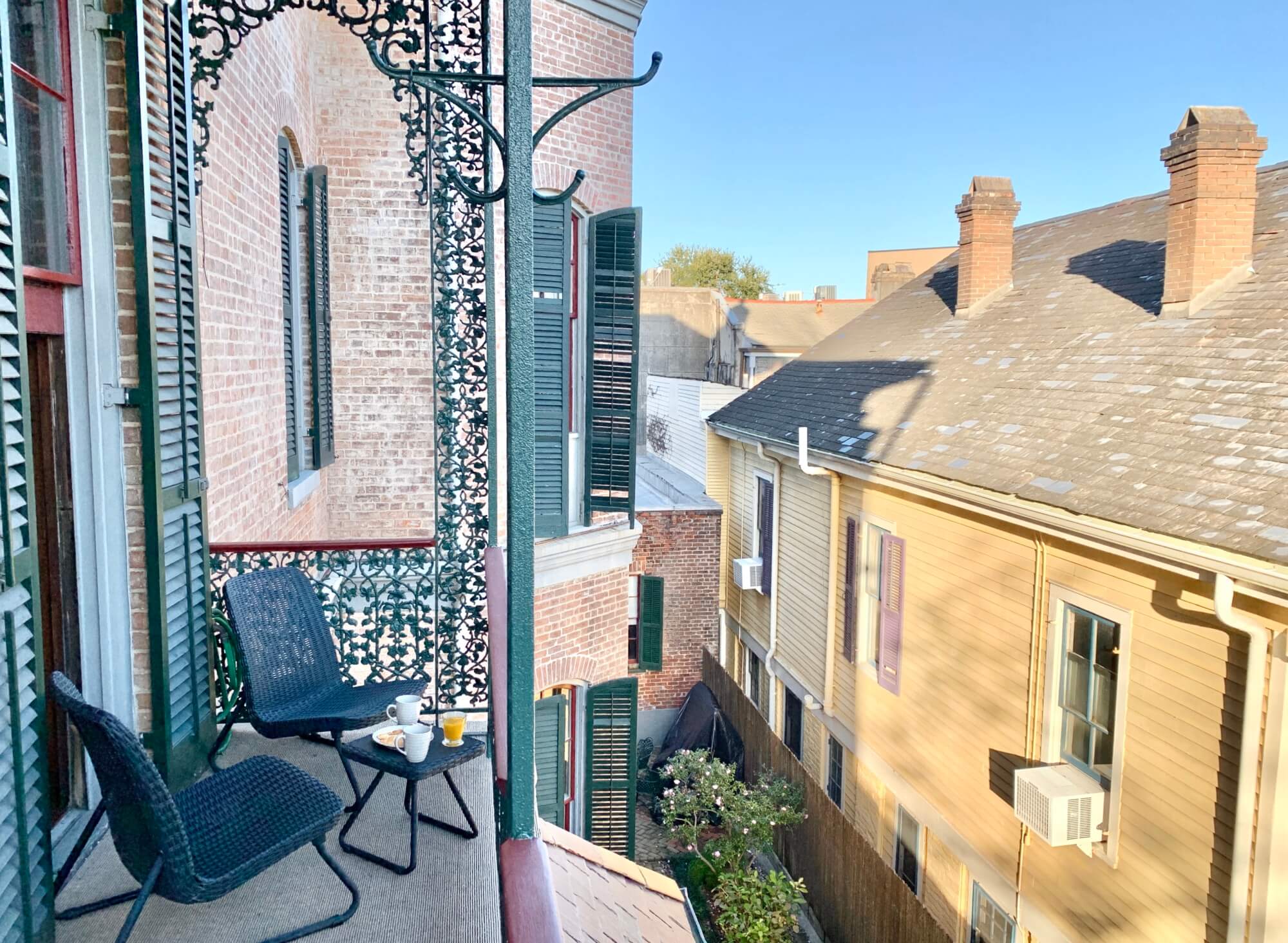 A small balcony with two chairs, a table set with breakfast, and a view of neighboring buildings.