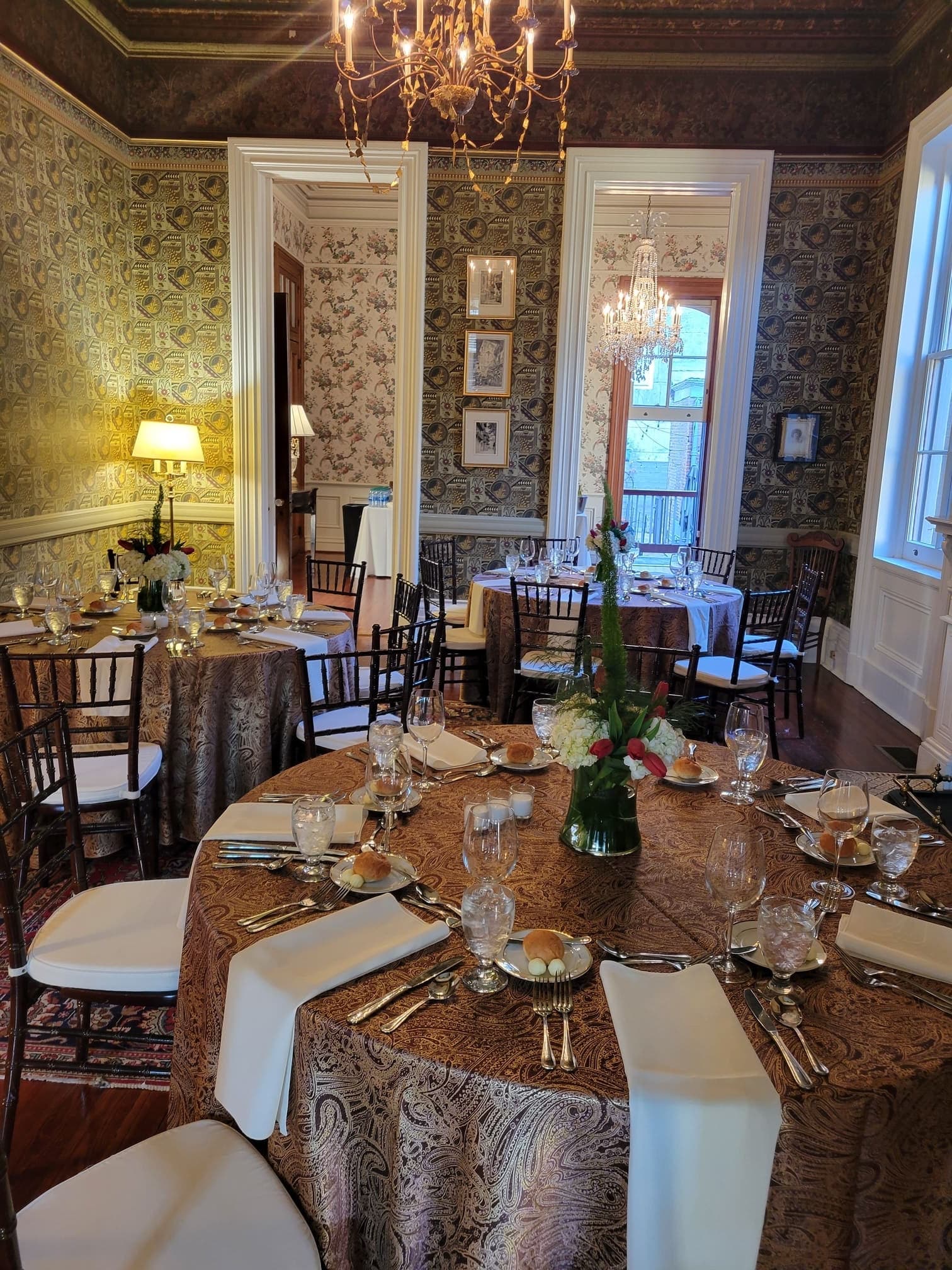 An elegantly set dining room featuring patterned wallpaper, ornate chandeliers, and tables arranged for a formal meal.