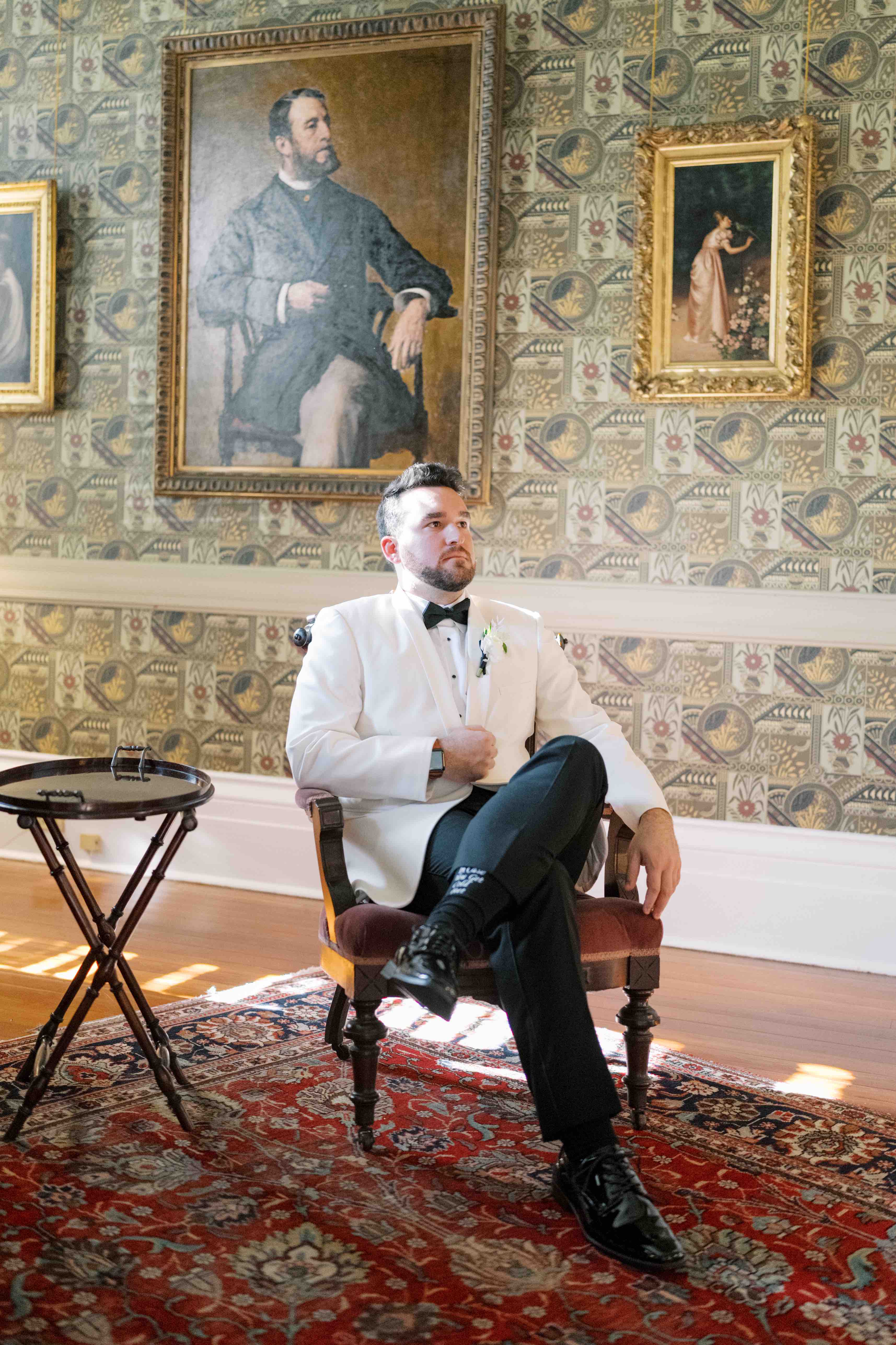 A man in a white tuxedo sits thoughtfully in an ornate room with vintage artwork.