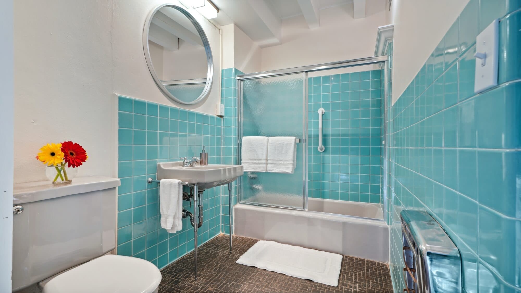 A bright bathroom featuring turquoise tiled walls, a glass shower, and a small bouquet of flowers on the counter.