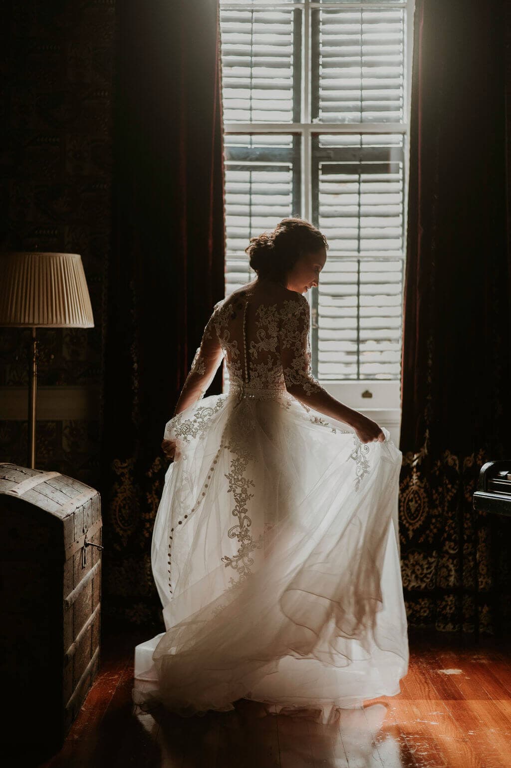 A bride in a flowing white gown stands by a window, illuminated by soft light.