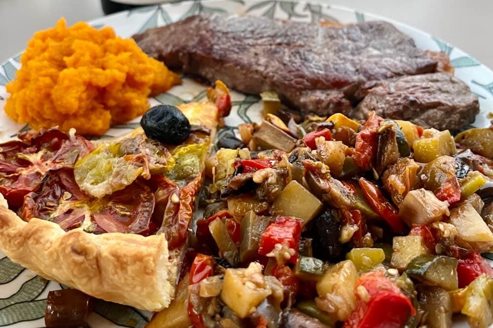 A plate featuring a piece of steak, a serving of mashed sweet potatoes, and a colorful vegetable medley with a slice of savory pie.