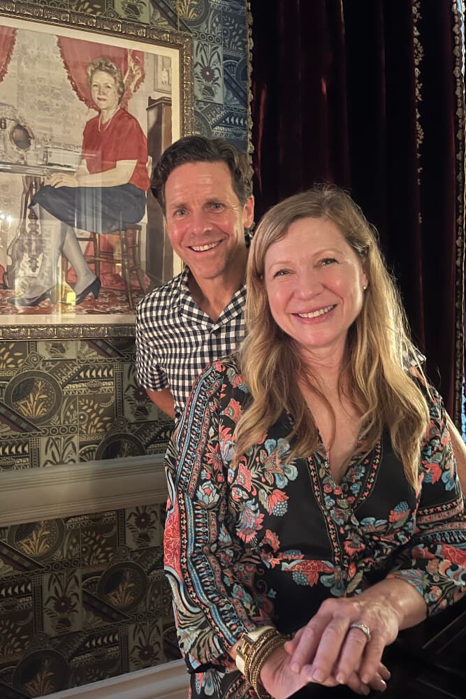 A man and a woman smile while standing in front of a vintage portrait on a patterned wall.