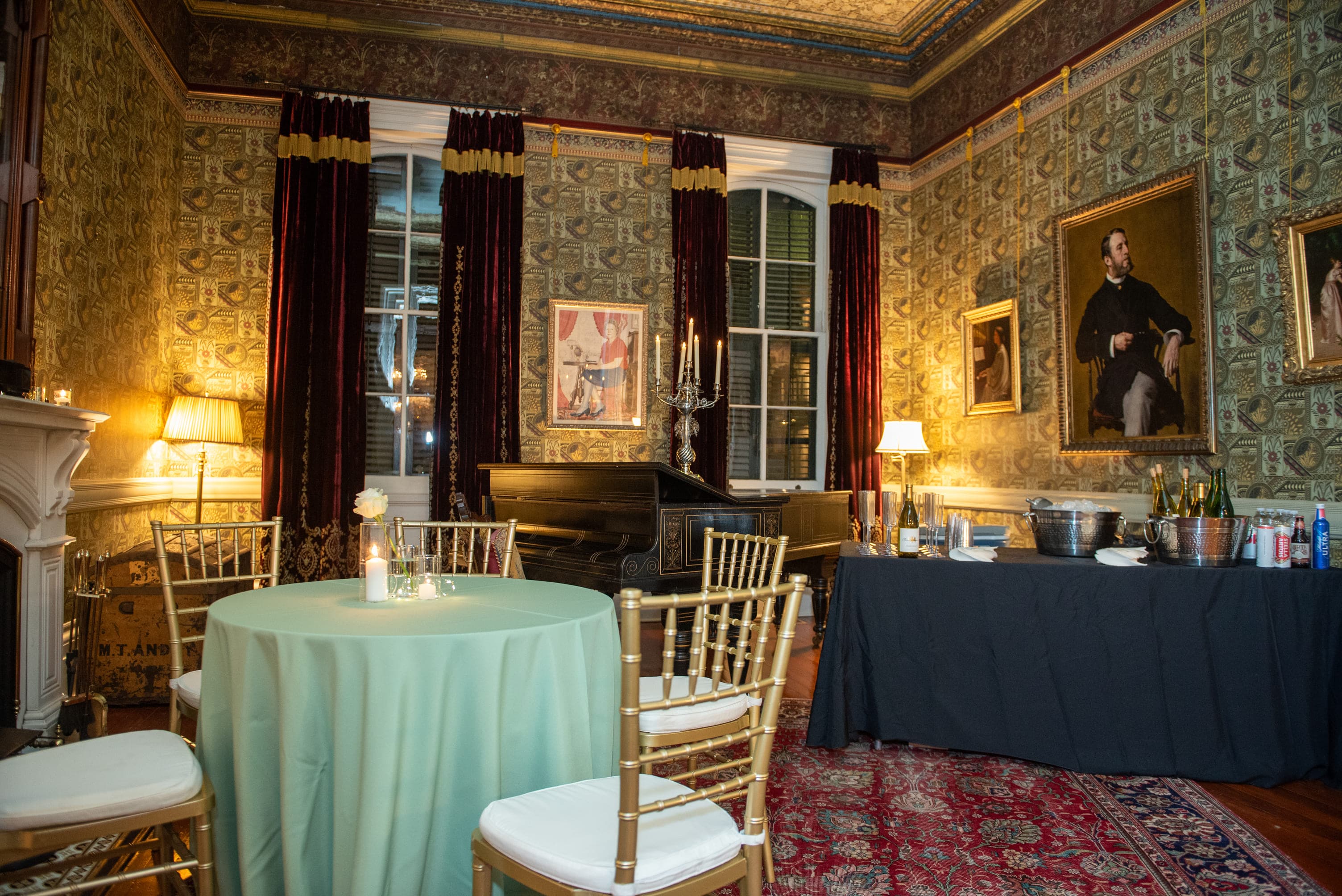 An elegantly decorated room featuring a green table, gold chairs, and art, with a buffet setup against ornate wallpaper.
