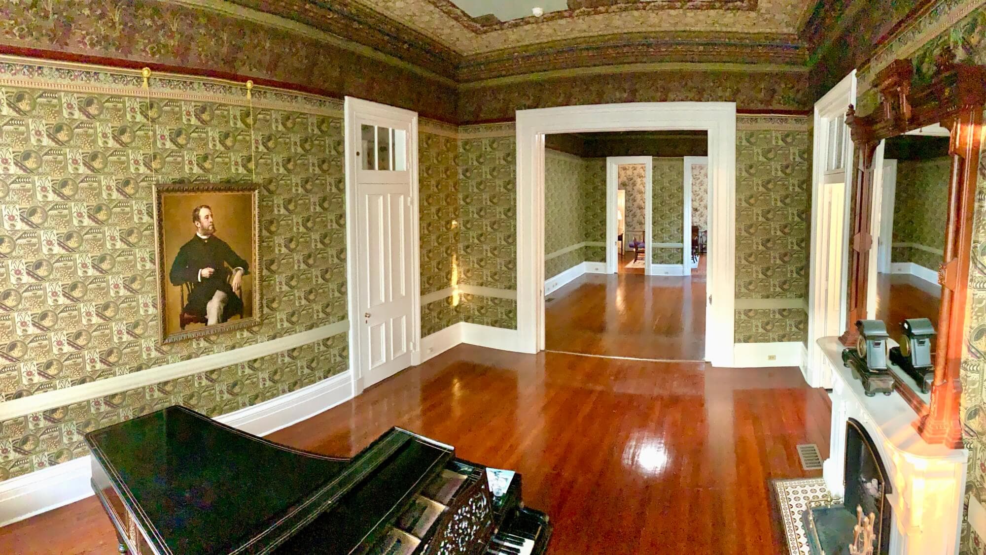 A spacious, vintage room featuring a grand piano, ornate wallpaper, and a framed portrait.