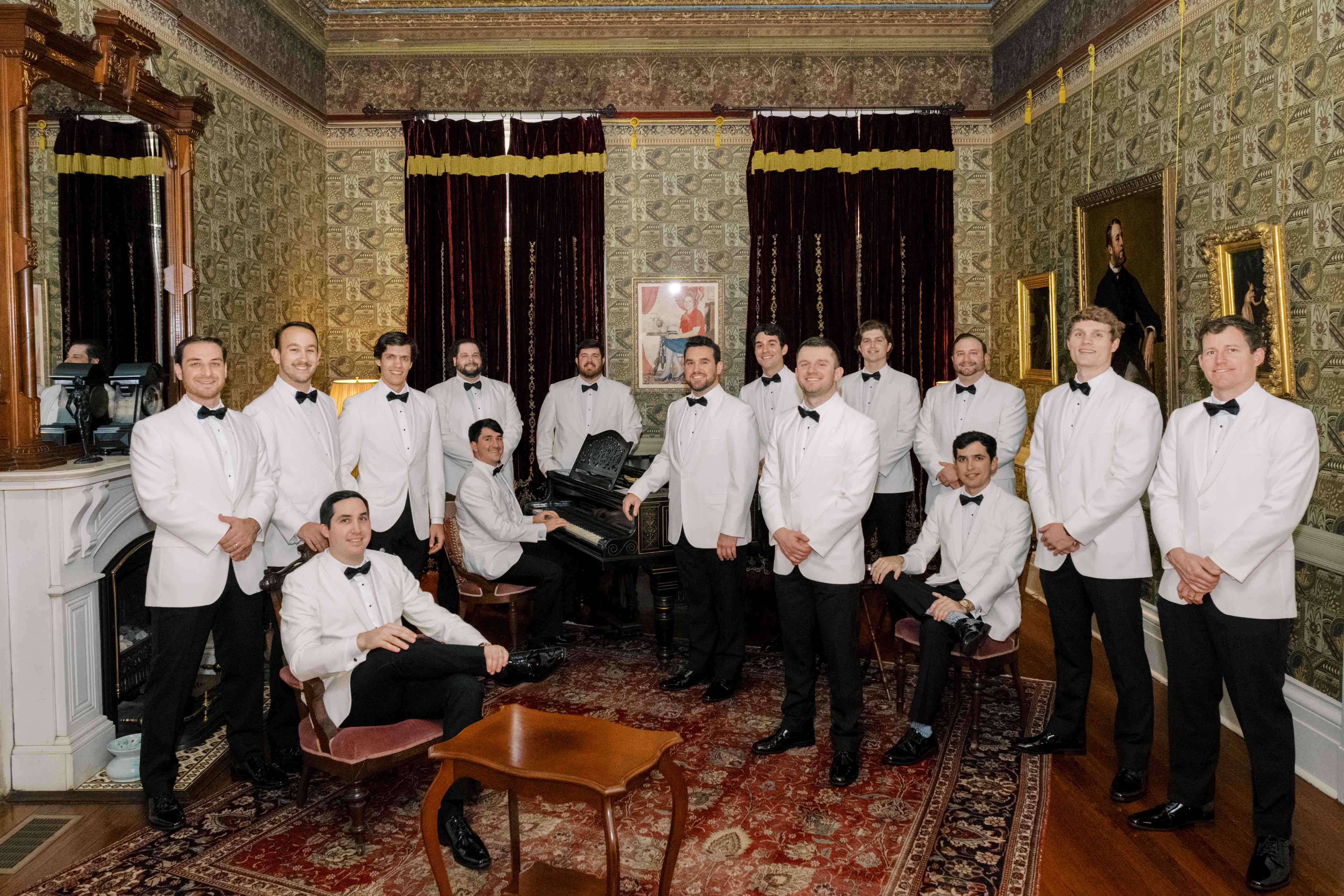 A group of men in white tuxedos pose elegantly in a richly decorated room with a piano.
