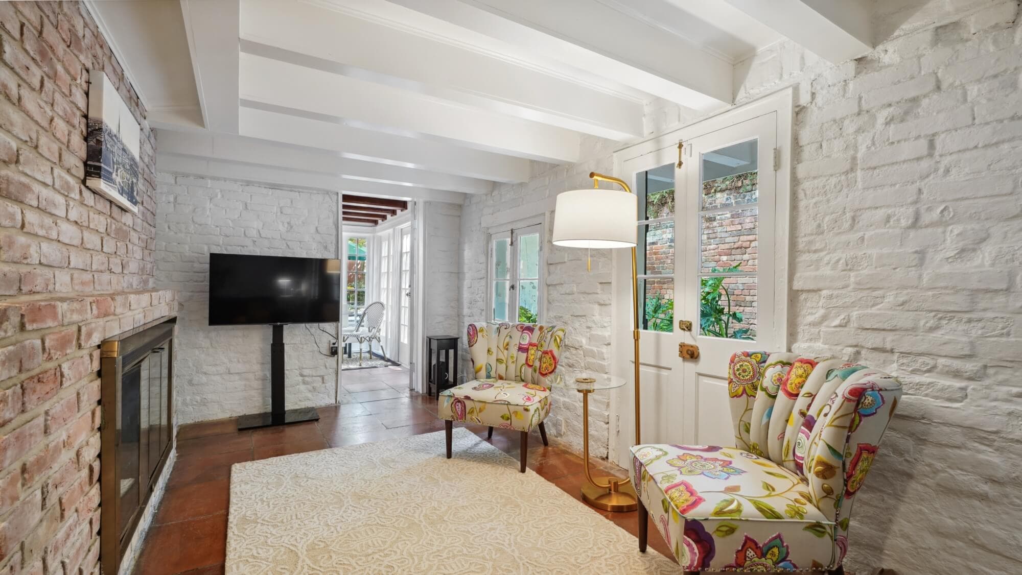 A cozy living room featuring floral armchairs, a brick wall, and a flat-screen TV.