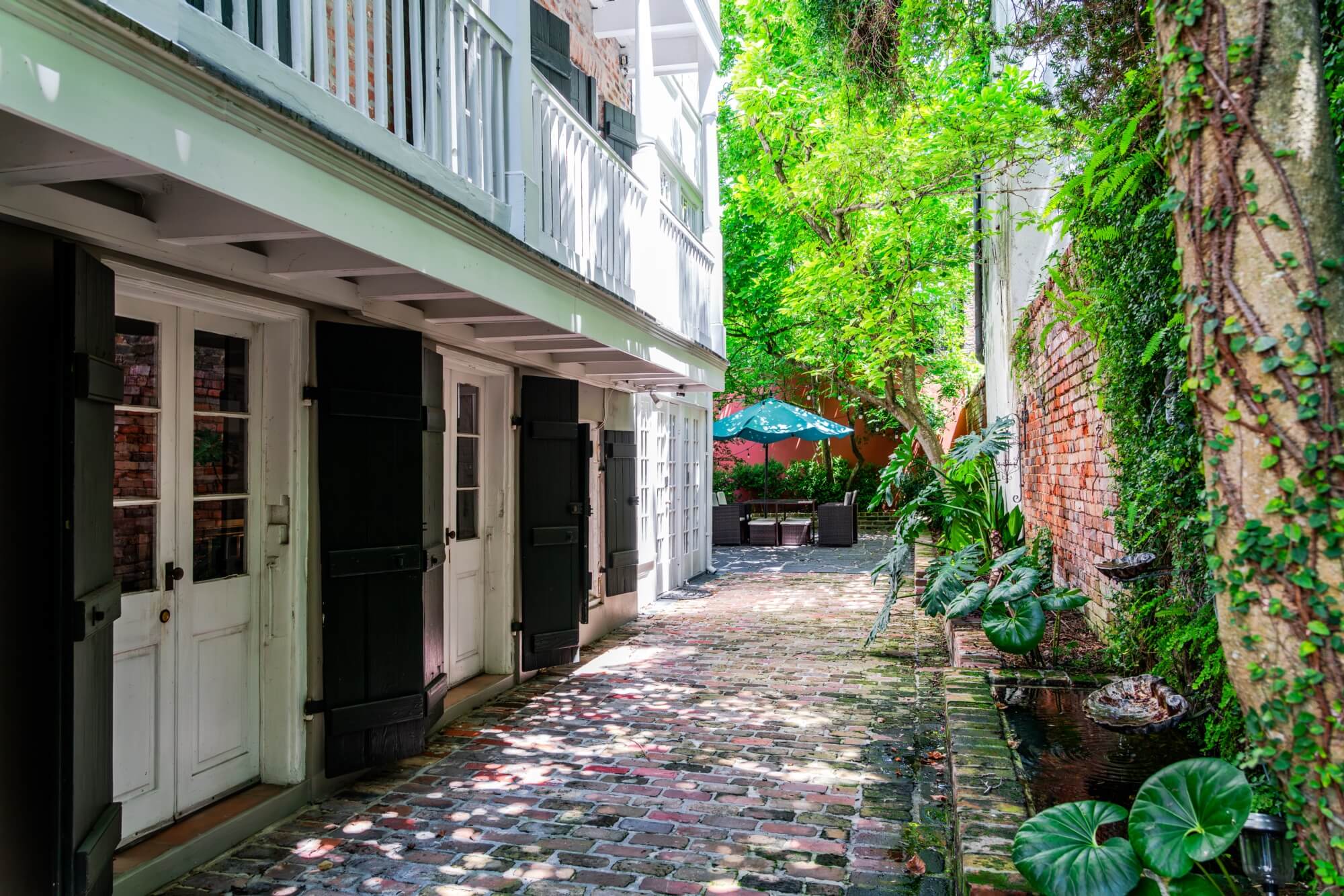 A narrow, cobblestone alleyway lined with greenery and doorways, leading to a cozy outdoor seating area.