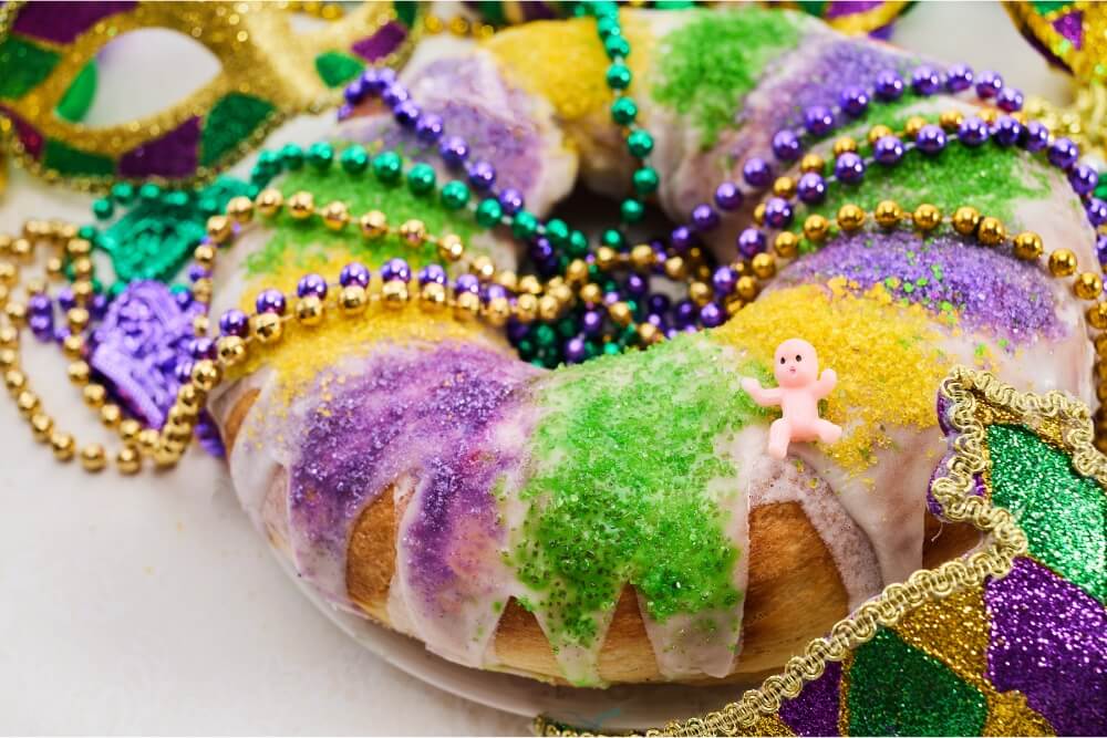A colorful king cake decorated with sprinkles and beads, featuring a small plastic figurine.