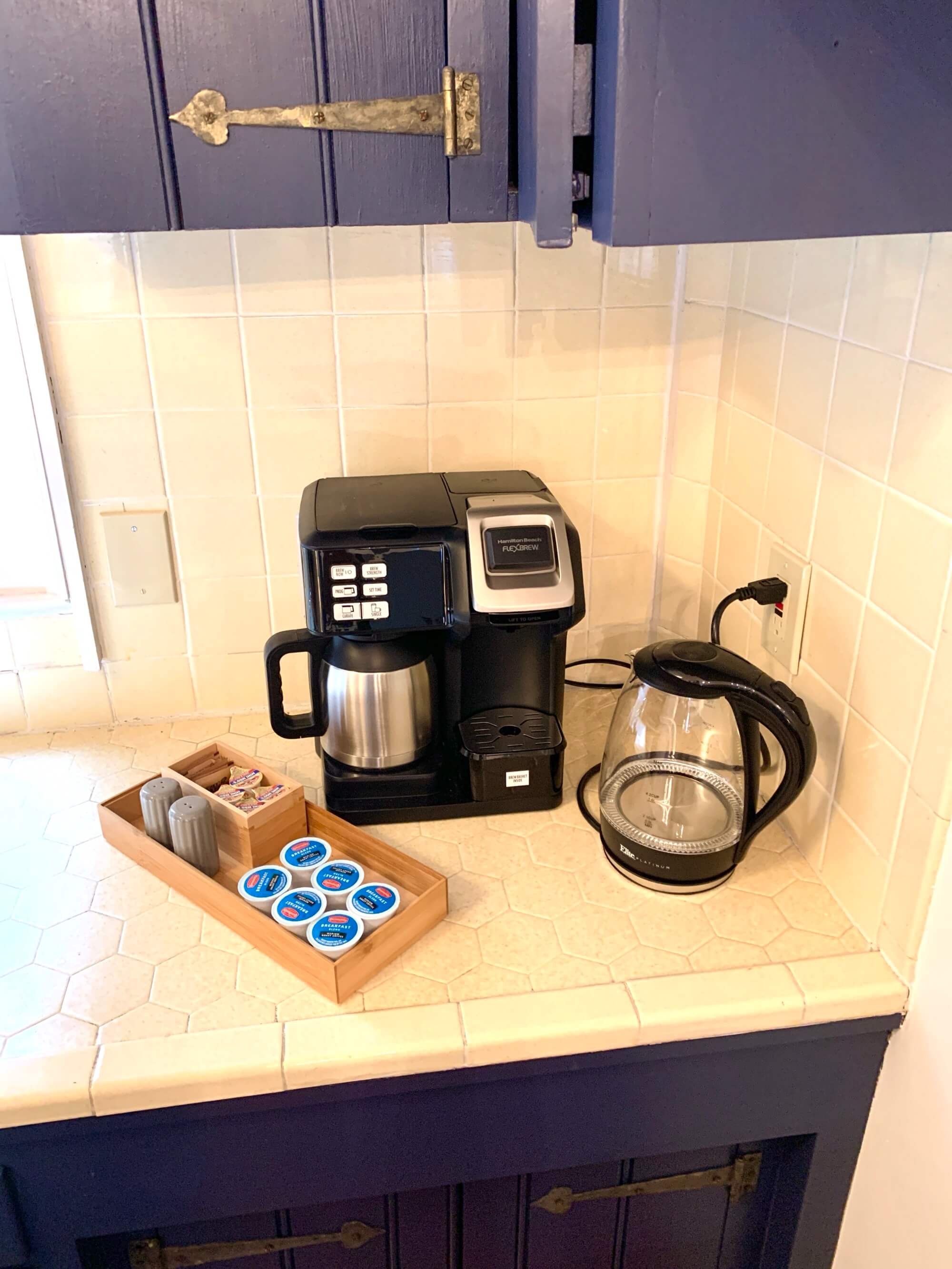 A coffee maker and kettle on a countertop with a tray of coffee pods and utensils.