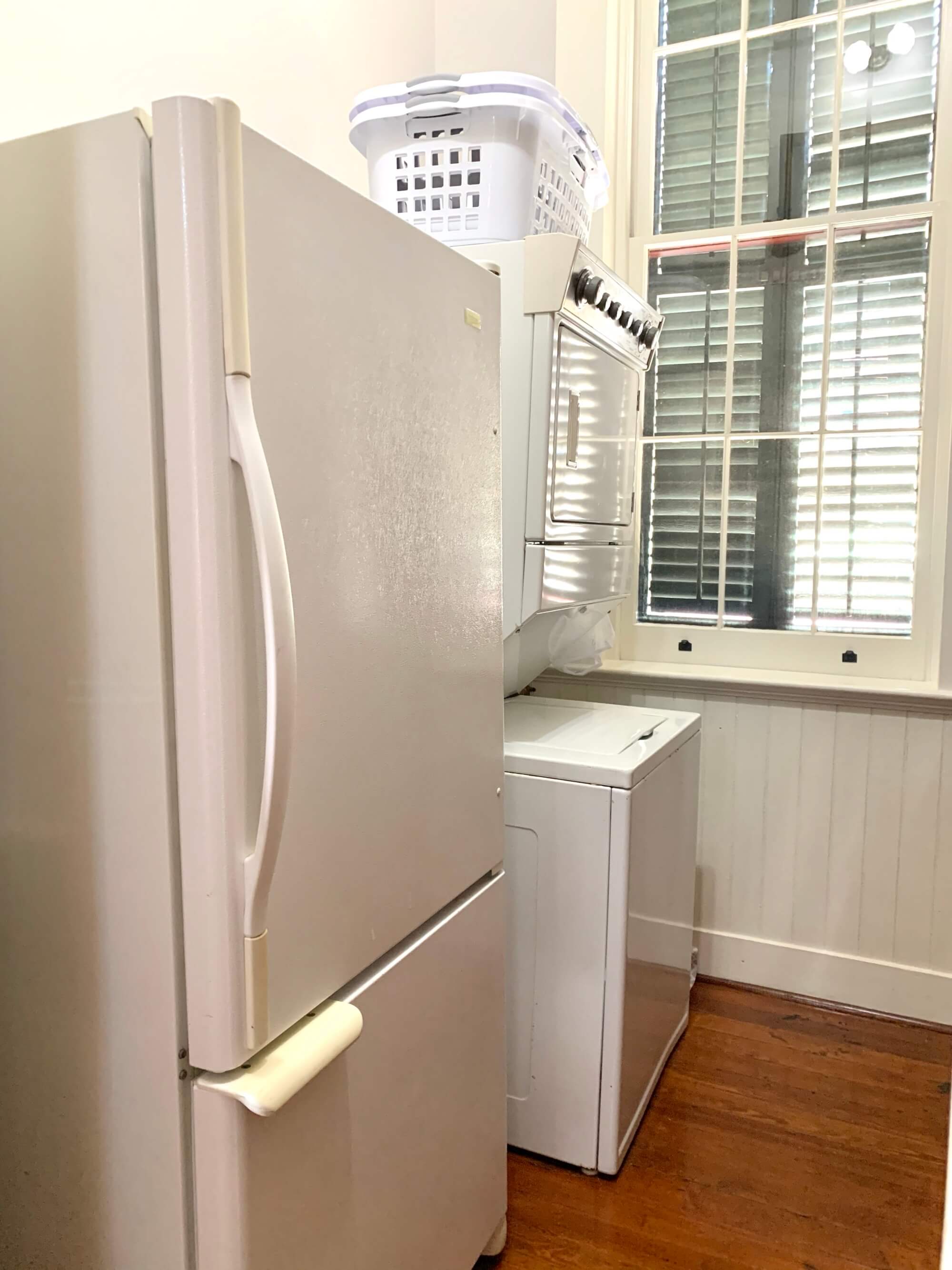 A white refrigerator and a stacked washer-dryer are positioned next to a window with shutters.