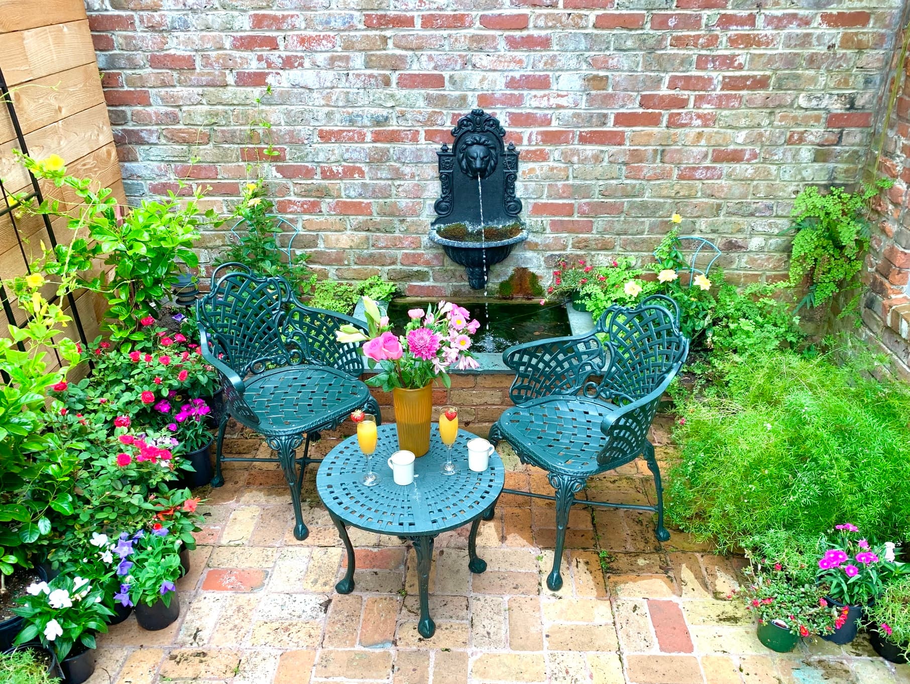 A cozy garden nook featuring two green chairs, a small table with drinks, and a brick wall adorned with plants and a fountain.