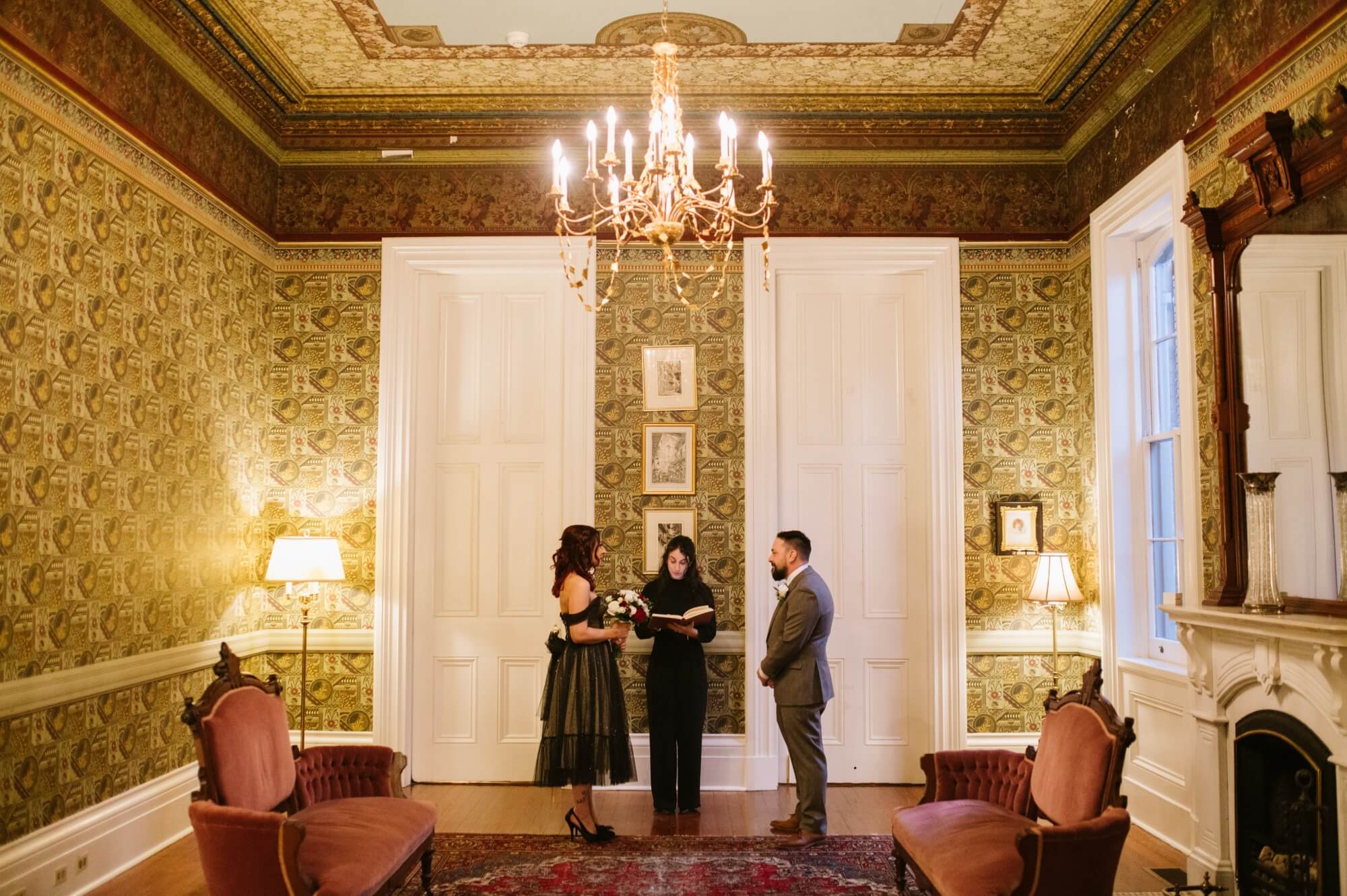 A couple stands together for a ceremony in an elegantly decorated room with vintage wallpaper and a chandelier.