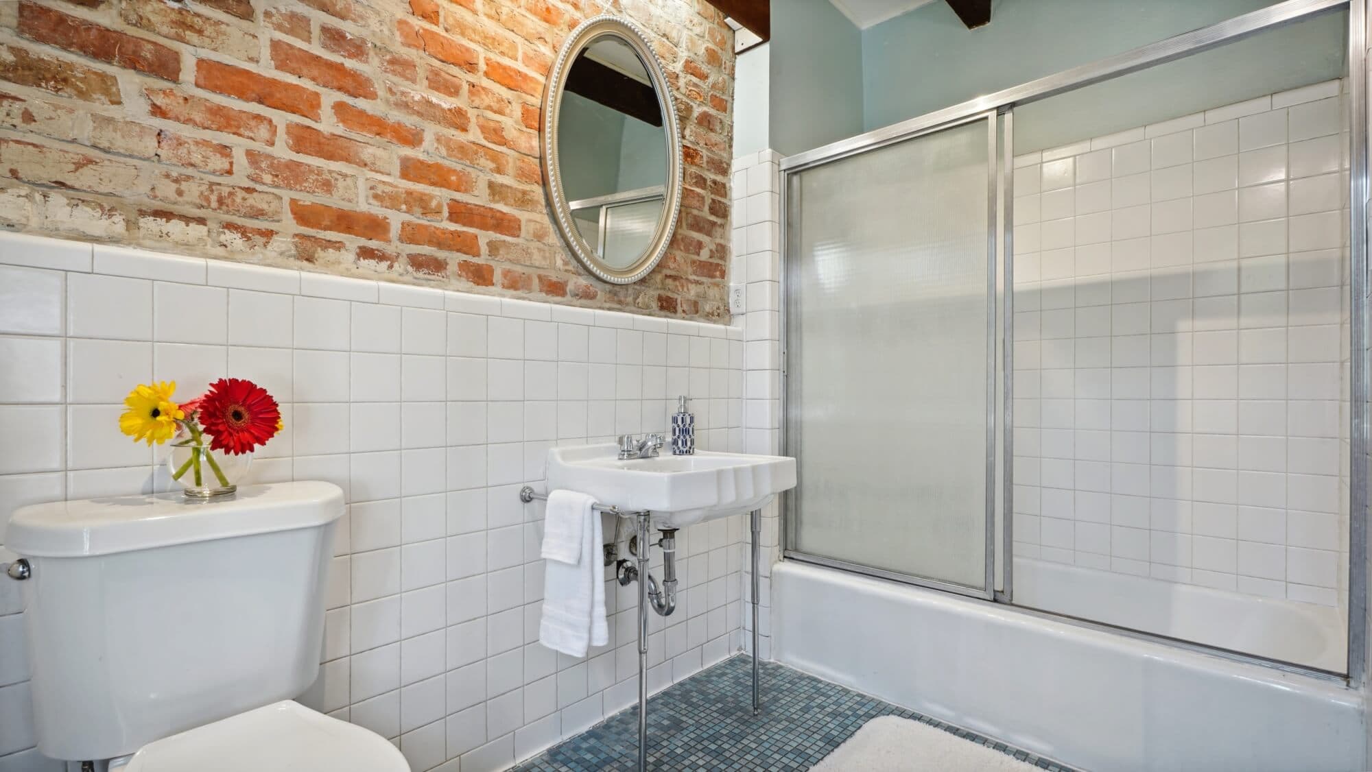 A modern bathroom featuring brick walls, a round mirror, a toilet, a sink with flowers, and a glass shower.