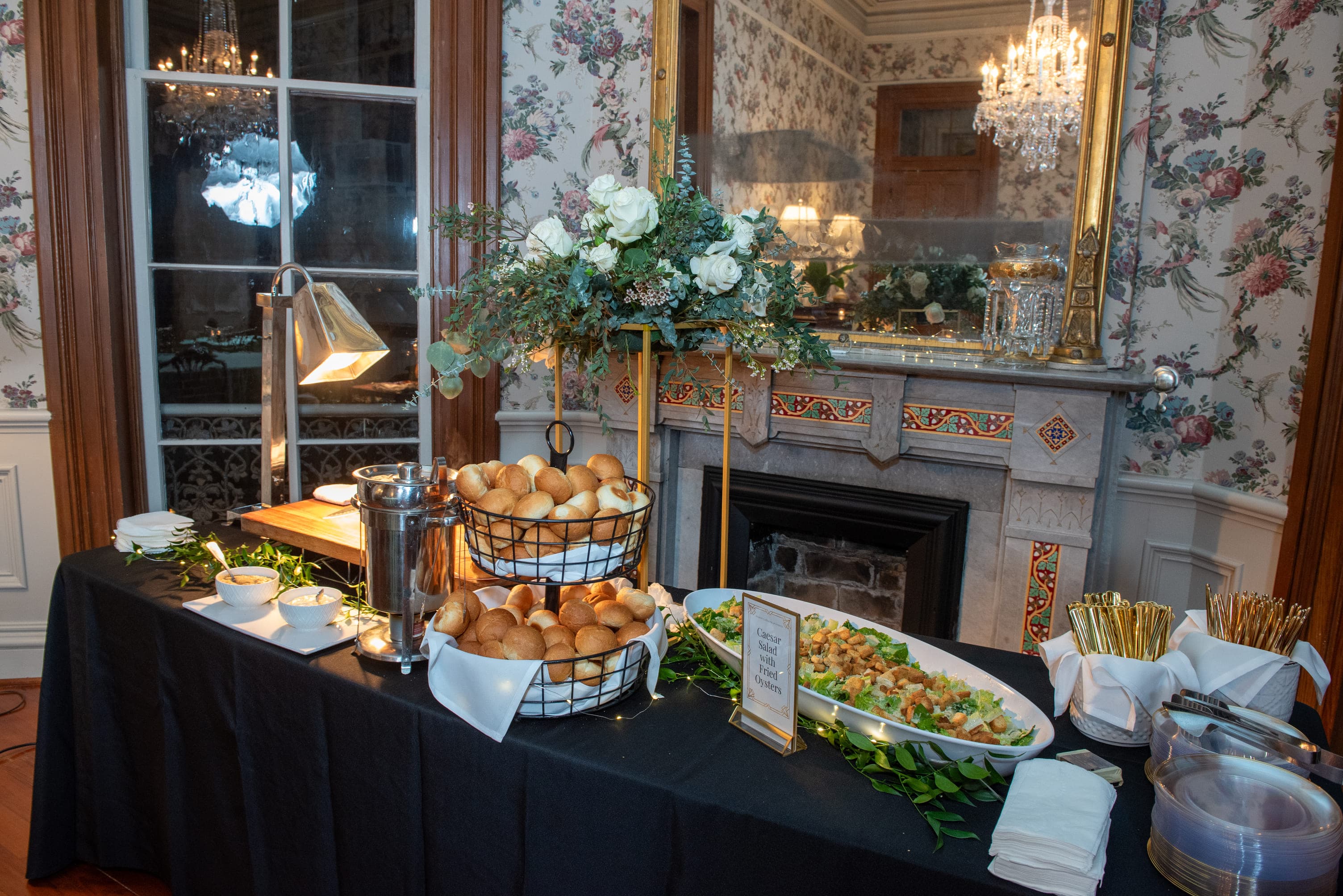 A elegantly arranged buffet table featuring rolls, a salad, and condiments, set against a floral wallpaper backdrop.