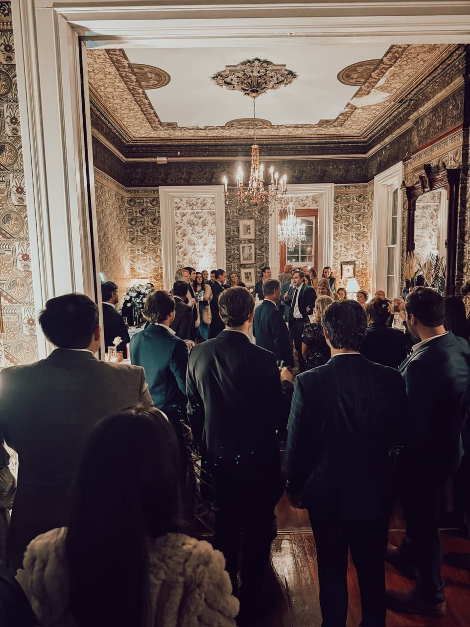 A crowd in formal attire gathers in a beautifully decorated room for a social event. A crowd in formal attire gathers in a beautifully decorated room for a social event.